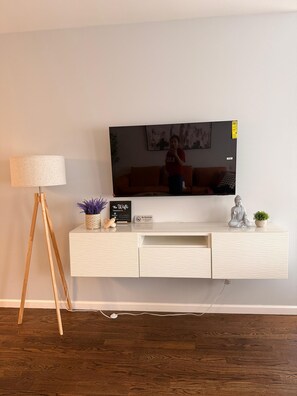 Una sala de estar moderna con un televisor de pantalla plana montado en una pared blanca, una lámpara de pie de madera, una consola blanca con objetos decorativos y un suelo de madera oscura.
