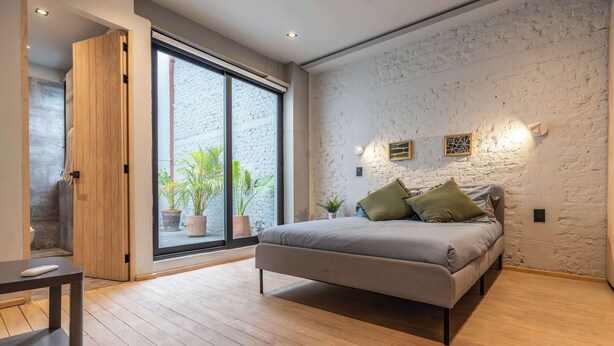 Une chambre avec un lit, un plancher en bois, une porte-fenêtre coulissante et un mur en briques blanches.