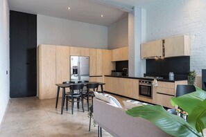 Eine moderne Küche mit Holzschränken, einem schwarzen Kühlschrank, einem Essbereich mit Tisch und Stühlen und einer großen Pflanze im Vordergrund.