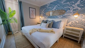 Ein ordentlich bezogenes Bett mit Kissen und Decke, eine Nachttischlampe, eine Wand mit Wellenmuster und ein Blumentopf.