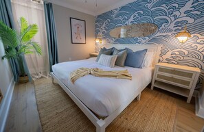 Ein ordentlich bezogenes Bett mit Kissen und Decke, eine Nachttischlampe, eine Wand mit Wellenmuster und ein Blumentopf.