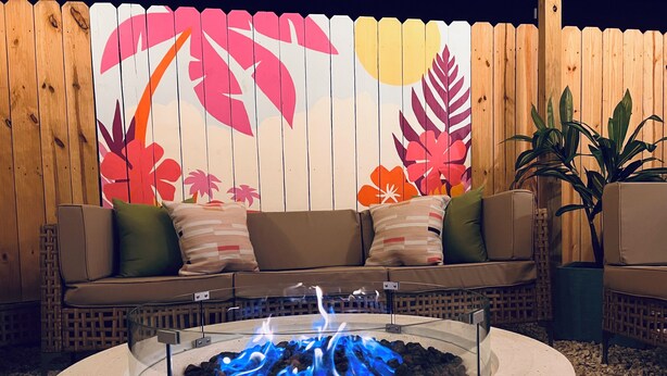 Ein runder Feuerkorb im Freien mit blauen Flammen, ein Holzzaun mit floralen Verzierungen und ein beiges Sofa mit Kissen.