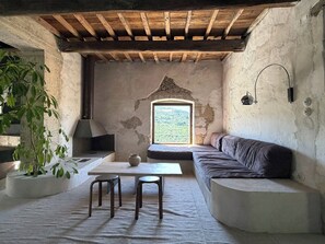 Ein rustikaler Raum mit Holzbalkendecke, einer Steinwand, einem großen Fenster, einem Sofa, einem Tisch, zwei Hocker und einem Blumentopf.