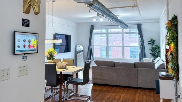 Ein modernes Wohnzimmer mit einer Couch, einem Esstisch mit Stühlen, einem Fernseher und einem großen Fenster mit Vorhängen.