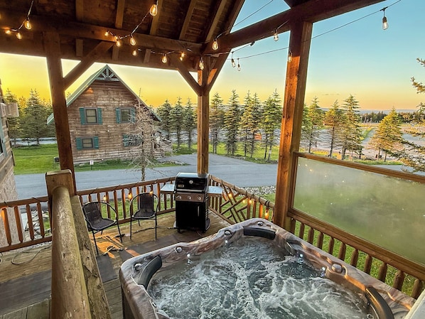 A wooden deck with a hot tub, outdoor seating, and a view of a cabin and lake.