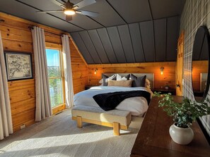 Un dormitorio con cama, mesitas de noche, ventilador de techo, ventana con cortinas y una planta.