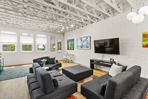 A modern living room with a white brick wall, large windows, a flat-screen TV, and a grey sectional sofa. - Luxury 2BR Condo on Peters St - Walk to FIFA & Mercedes-Benz Stadium (Atlanta)