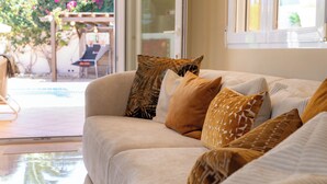 Un sofá color arena con cojines estampados, una puerta de cristal que da a un espacio exterior con una silla de patio, y una ventana con persianas.