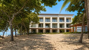 Un hotel frente a la playa con una hilera de habitaciones, un área de estar al aire libre techada y una playa de arena.