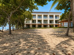 Un hotel a peu de platja, amb una filera d'habitacions, una zona de seients coberta a l'aire lliure i una platja de sorra.