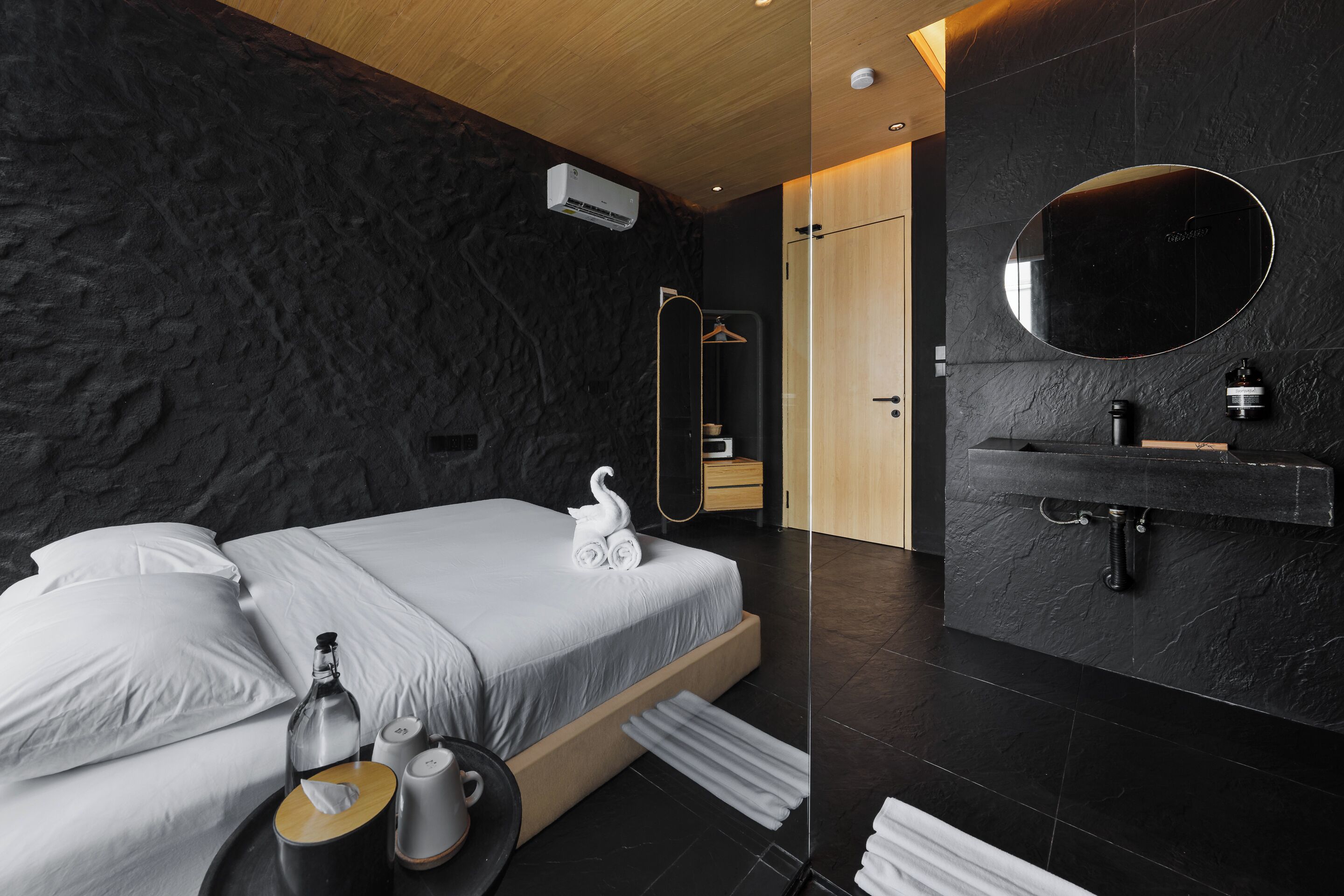 A modern bedroom with a stone-textured wall, a wooden ceiling, a bed with white linens, a bedside table with a bottle, a cup, and a tissue box.