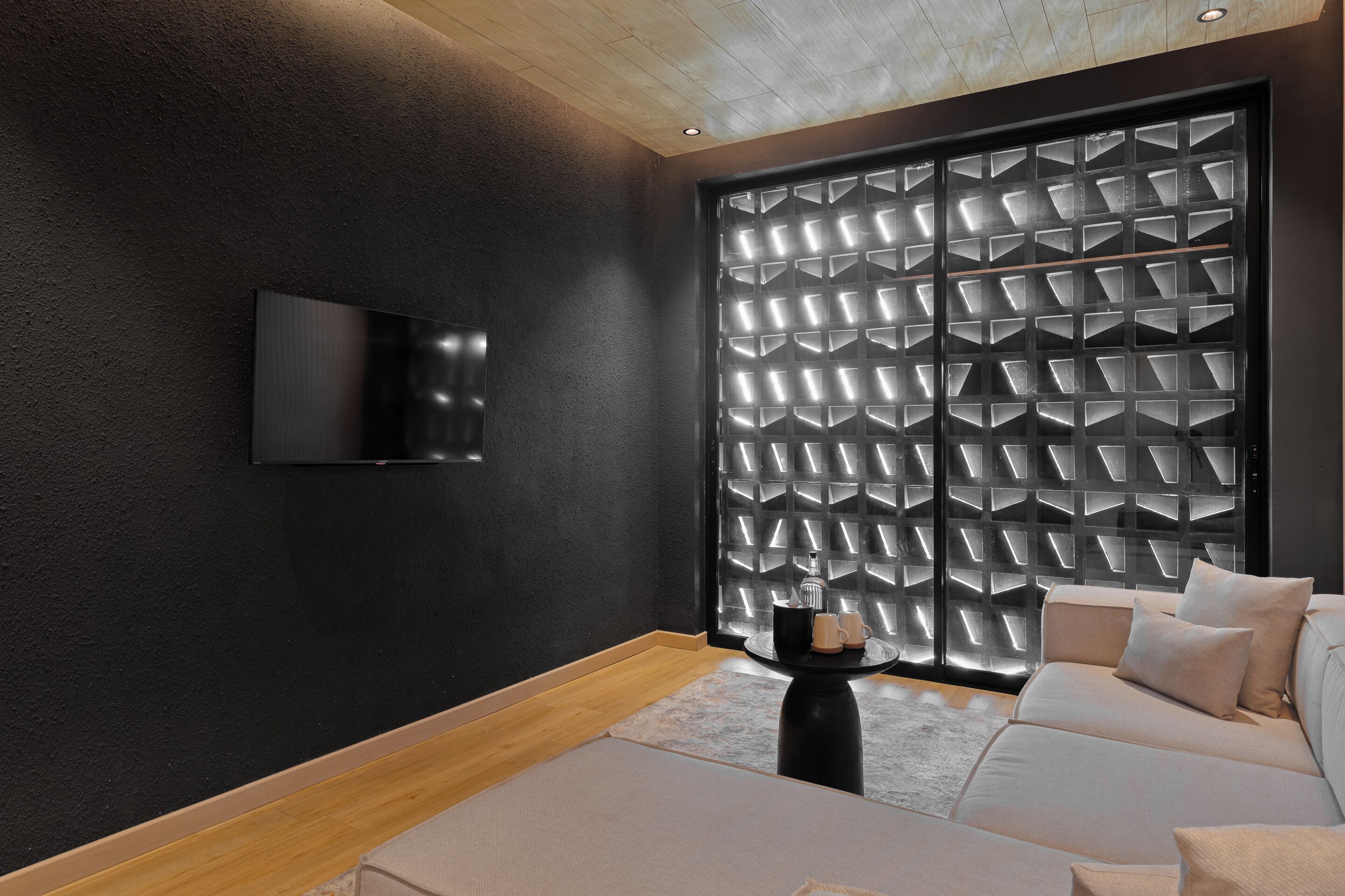 A modern room with a black textured wall, a mounted TV, a patterned sliding door, and a beige sofa with pillows.
