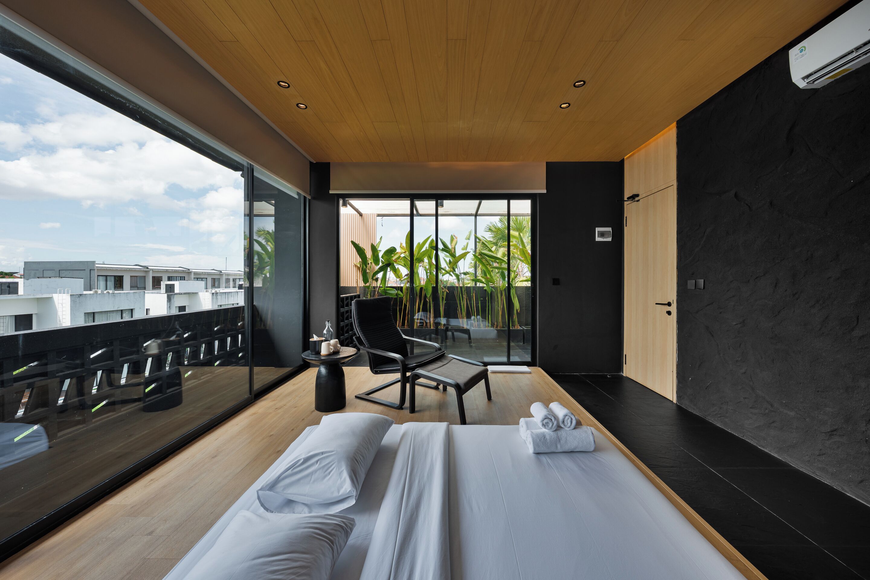 Une chambre d’hôtel moderne dotée d’un grand lit, d’un coin salon avec un fauteuil et une petite table, et offrant une vue sur la ville grâce à de grandes fenêtres.