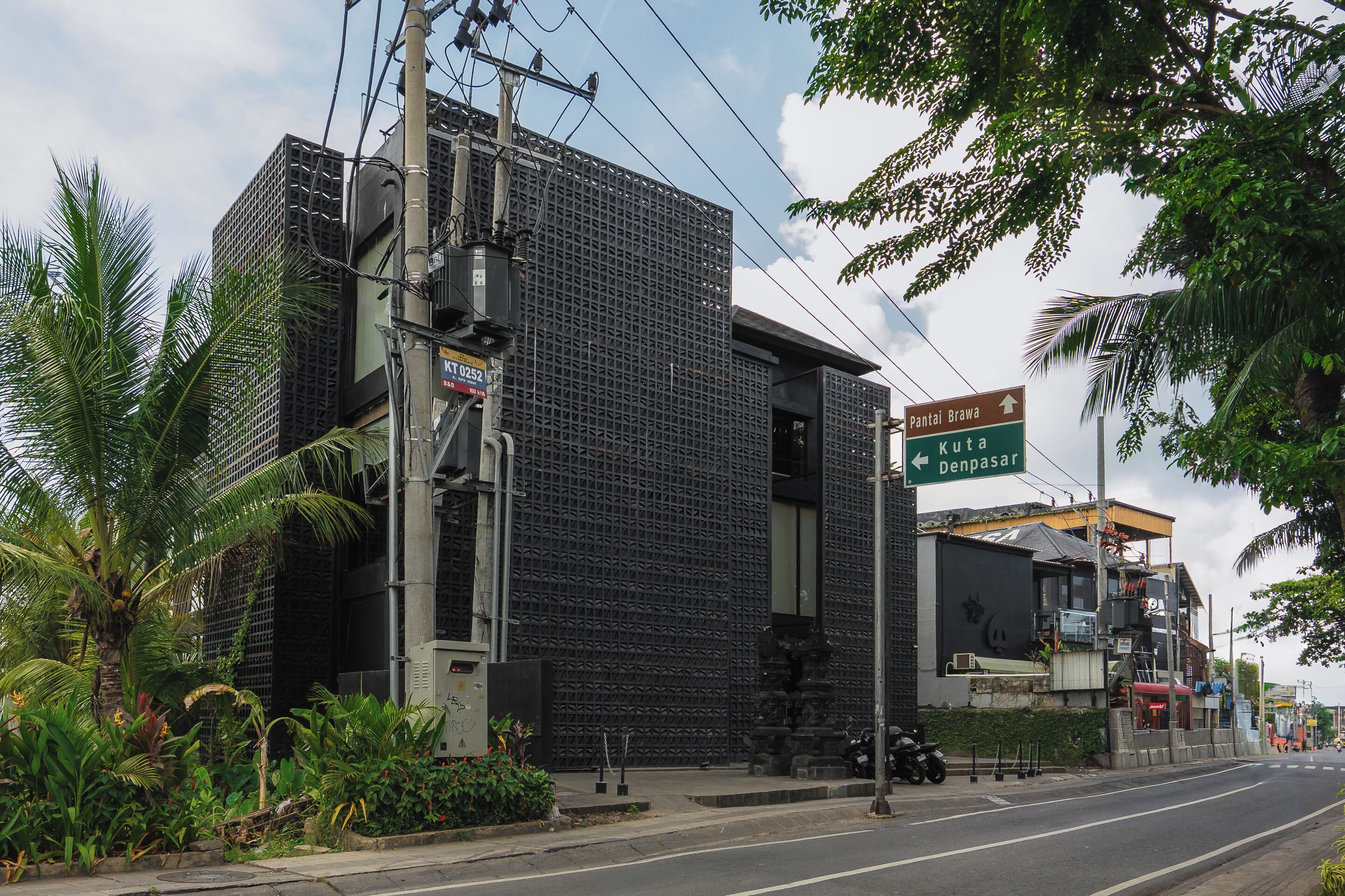A modern building with a grid-like facade, a street sign indicating directions to 'Pantai Brawa' and 'Kuta Denpasar,' and a motorcycle parked on the sidewalk.