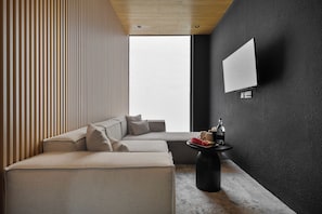 Un salon moderne avec un canapé d’angle, une petite table ronde sur laquelle se trouve un plateau de collations et un téléviseur à écran plat fixé à un mur foncé.