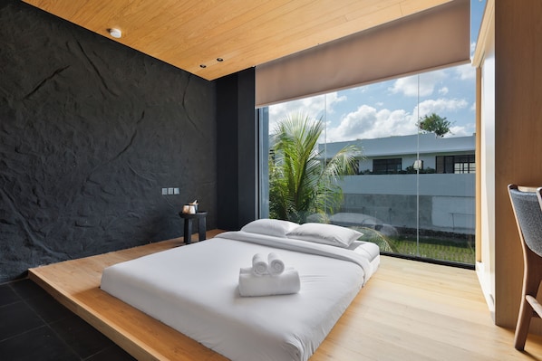 A modern bedroom with a large bed, a view of the sky and palm tree, and a wooden ceiling.