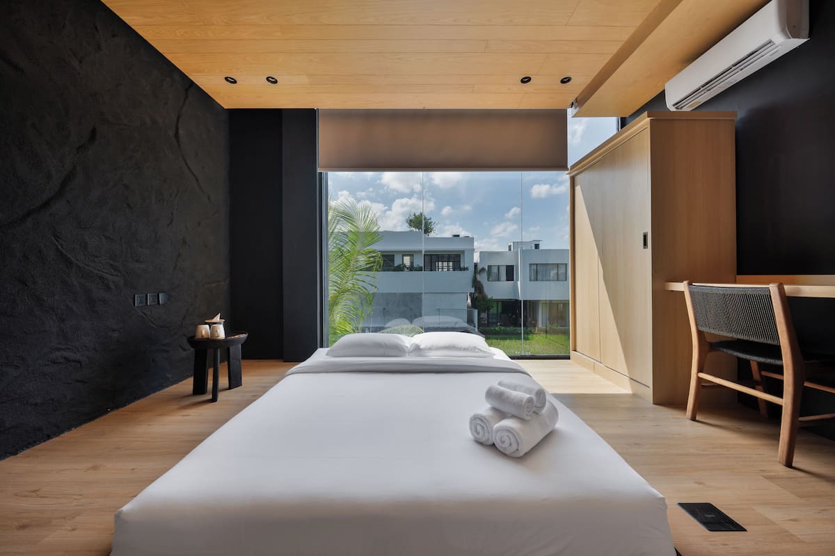 A modern hotel room with a large bed, a desk, and a view of buildings through a large window.