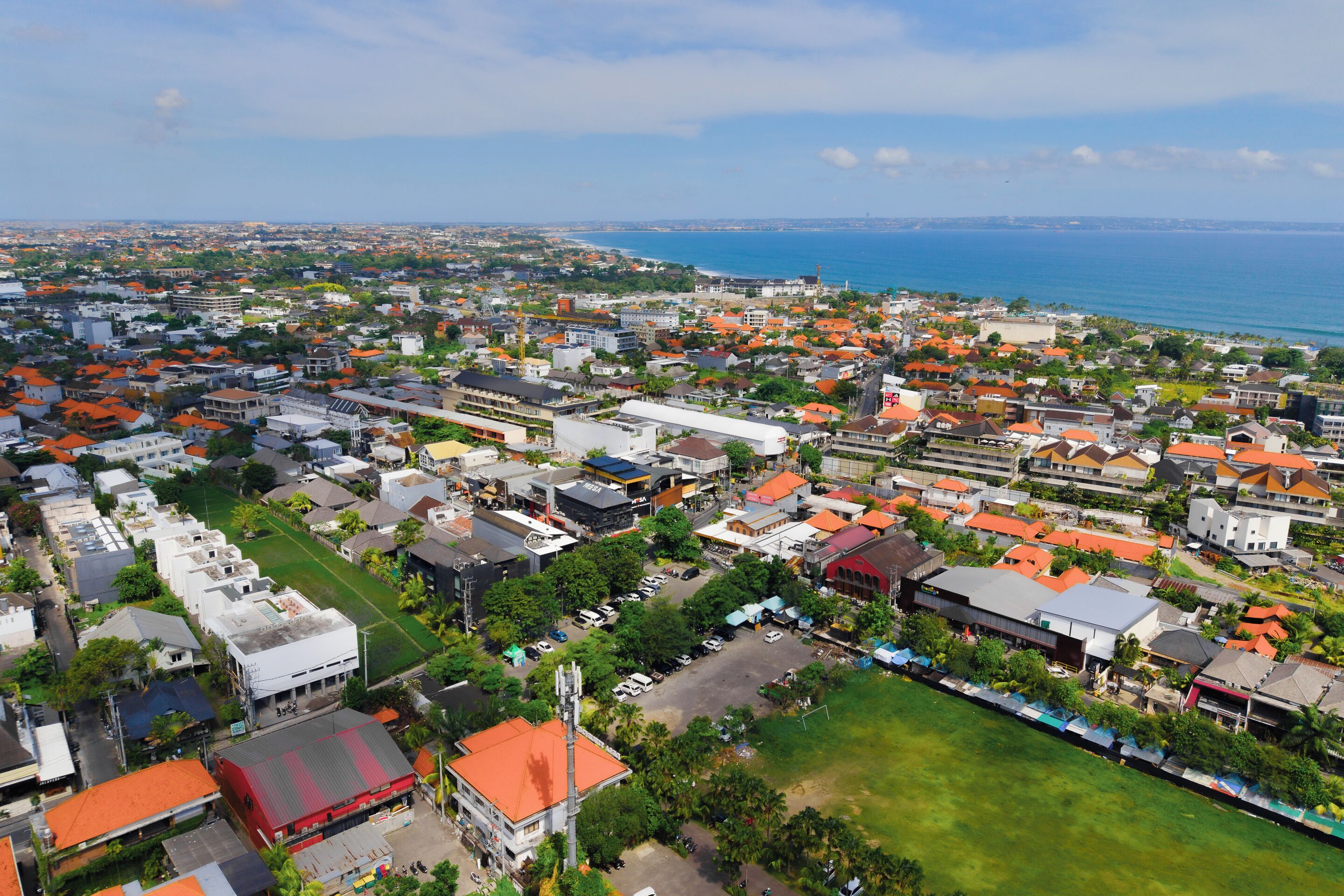 An aerial view of a coastal town with a mix of residential and commercial buildings, a prominent red-roofed building, and a view of the sea.