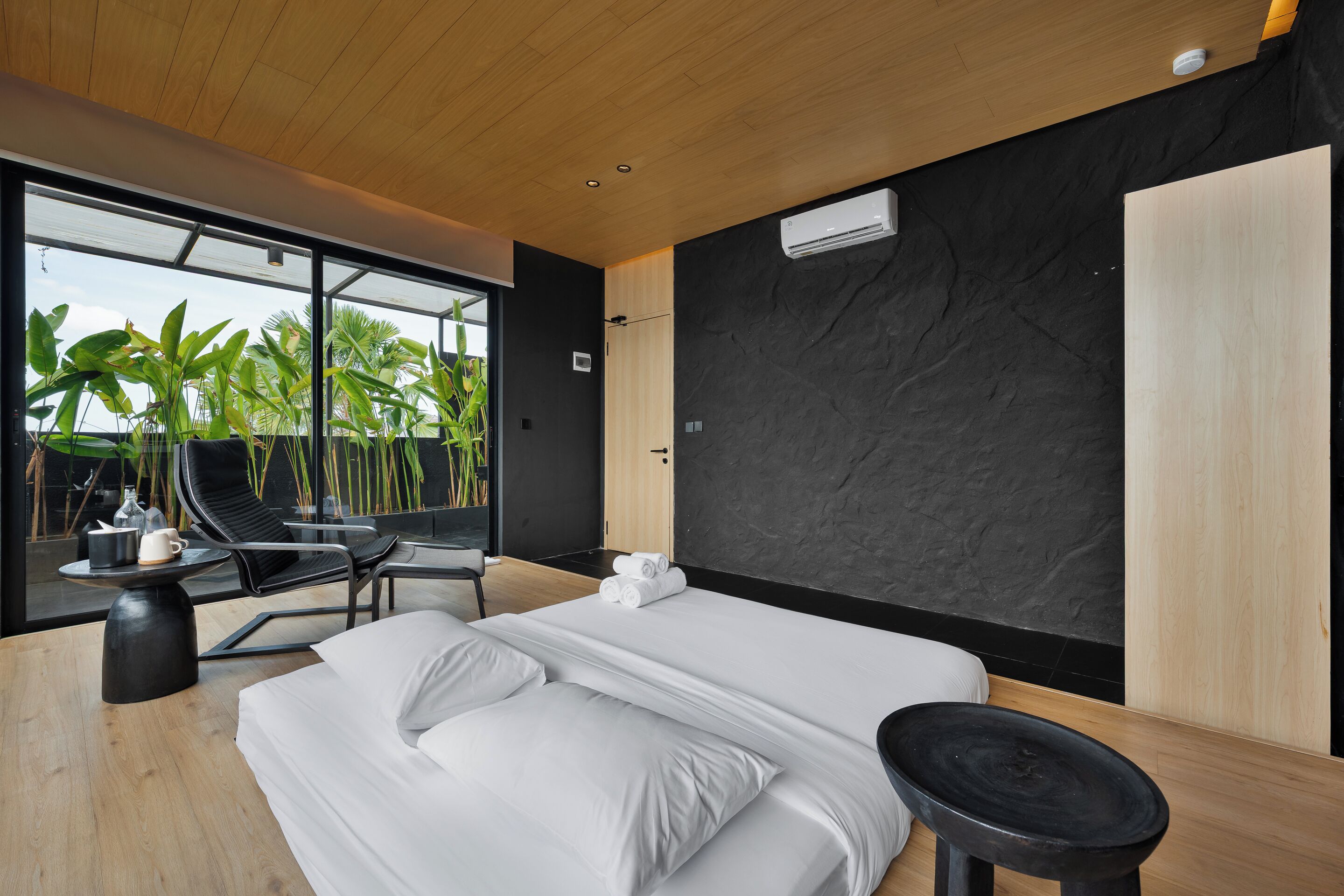A modern bedroom with a large bed, a black chair, a small table, and a view of palm trees through large windows.