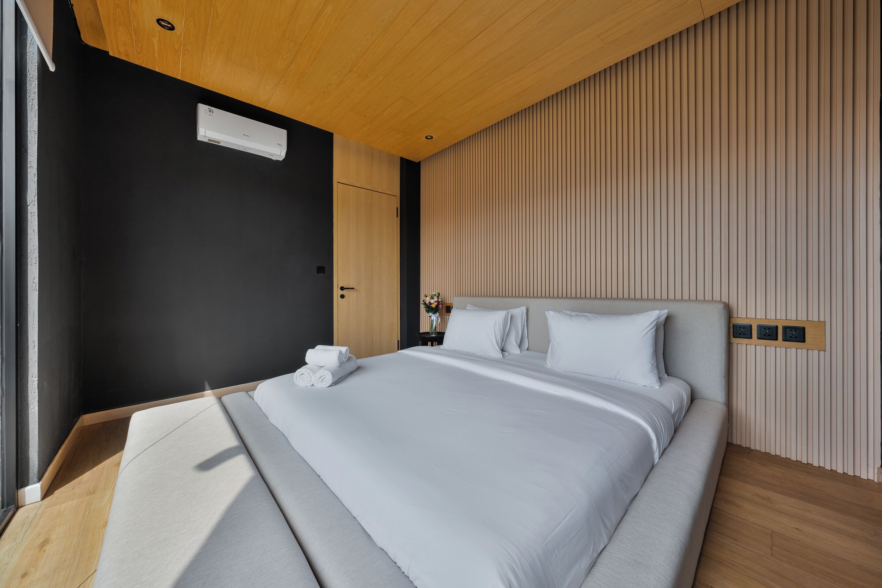 A modern hotel room with a large bed, wooden ceiling, and a wall-mounted air conditioner.