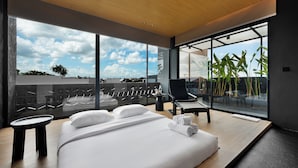 Una habitación de hotel moderna con una cama grande, un área de descanso con una silla y una mesa, y un balcón con plantas.