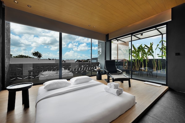 Une chambre d’hôtel moderne dotée d’un grand lit, d’un coin salon avec un fauteuil et une table, et d’un balcon agrémenté de plantes.