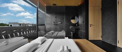 A modern hotel room with a large bed, a separate shower area, a wooden ceiling, and a large window offering a view of the sky and buildings.