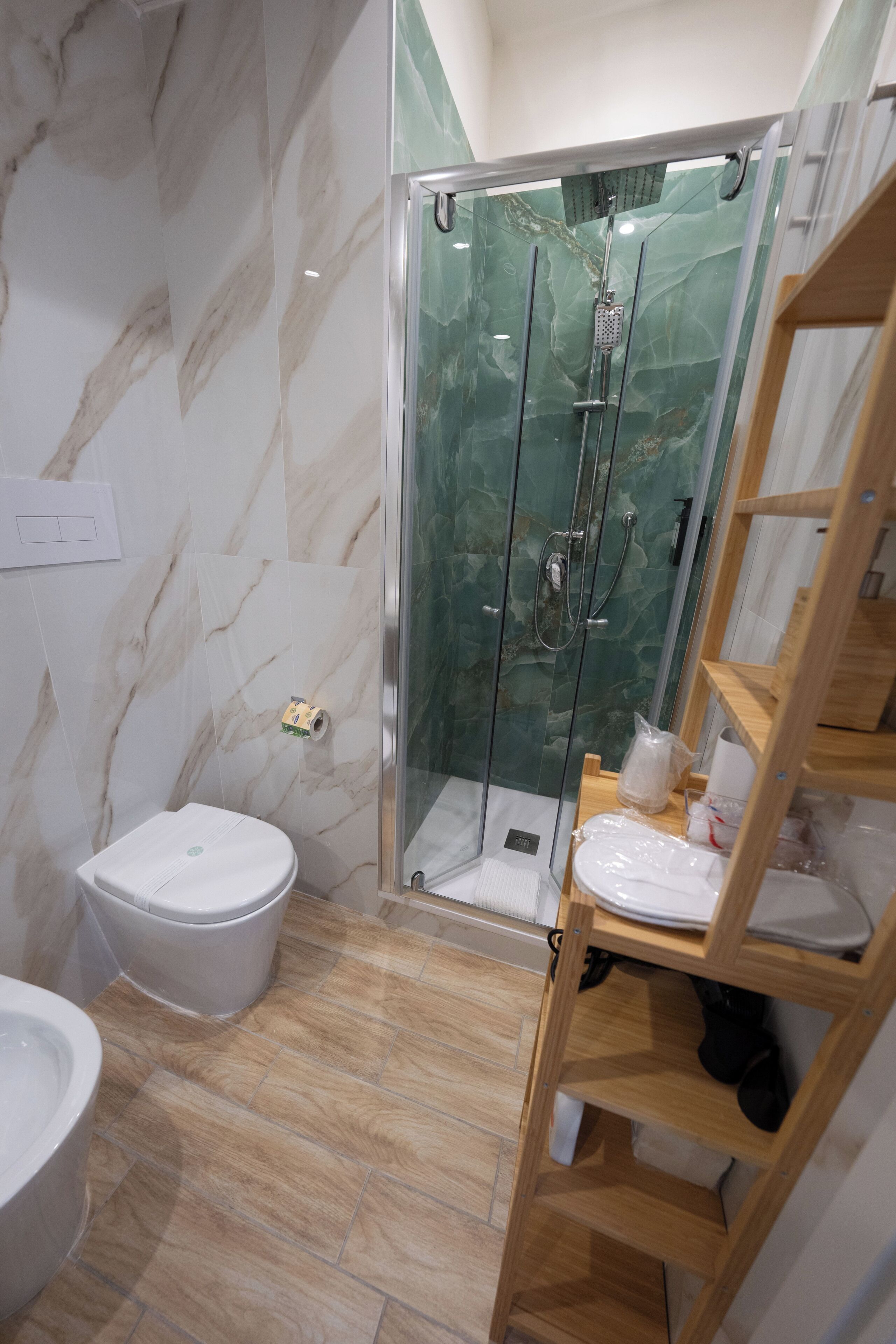 A bathroom with a marble wall, a glass-enclosed shower, a toilet, and a wooden shelf.