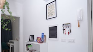 A room with a white wall, a shelf with books and a framed picture, a hanging plant, a desk, and a door. - Partesia  (Naples)