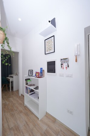 A room with a white wall, a shelf with books and a framed picture, a hanging plant, a desk, and a door. - Partesia (Naples)
