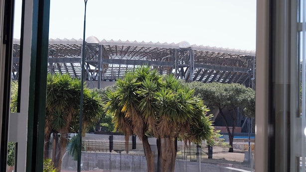 A window view of a stadium with a modern architectural design, palm trees, and a street lamp. - Partesia  (Naples)