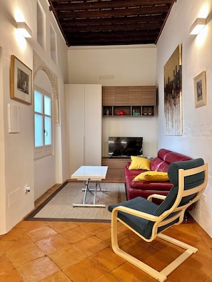 Ein Wohnzimmer mit einem roten Sofa, einem Holzstuhl, einem Fernseher und einer Küchenzeile im Hintergrund.
