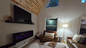 A cozy living room with a fireplace, a TV mounted on the wall, a comfortable sofa, and a lamp. - The A at Elm Creek - Modern Cabin Retreat ~ Hot tub, Game loft, EV charger (Hot Spring County)