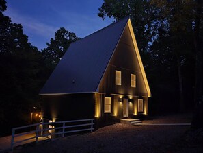 A large, dark-colored barn with a sloped roof, illuminated windows, and a white fence in front.