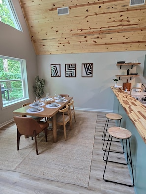 A dining area with a wooden table set for four, a bar with stools, and a view of greenery outside.