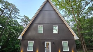 A dark-colored, triangular-roofed house with a front door, two windows on the ground floor, and a small porch area. - The A at Elm Creek - Modern Cabin Retreat ~ Hot tub, Game loft, EV charger (Hot Spring County)
