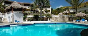 A swimming pool with lounge chairs, a thatched-roof structure, and a multi-story building with balconies.