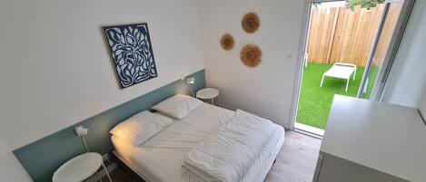 Un dormitorio con cama, mesitas de noche, lámpara empotrada en la pared, una obra de arte enmarco y vistas a un área verde exterior a través de una puerta corredera.