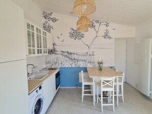 A kitchen with a washing machine, a dining table with chairs, and a wall mural. - LA NOUVELLE VAGUE 100M FROM THE SEA (La Guérinière)