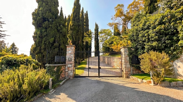 Una entrada con puerta de reja, pilares de piedra, camino de grava y diversidad de árboles y arbustos.