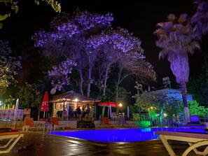 A nighttime outdoor pool area with a bar and seating, surrounded by trees and illuminated by purple lighting.