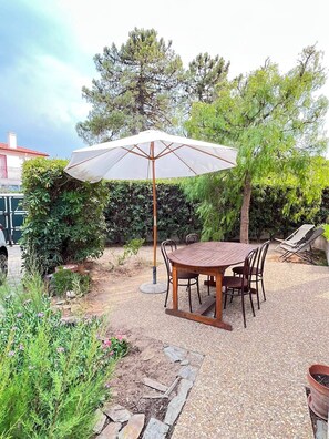 Eine Terrasse im Freien mit Tisch und Stühlen, umgeben von Pflanzen und einem großen Sonnenschirm.