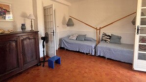 Una habitación con dos camas, un taburete pequeño azul, un armario de madera, un ventilador y una puerta que da a otra habitación.