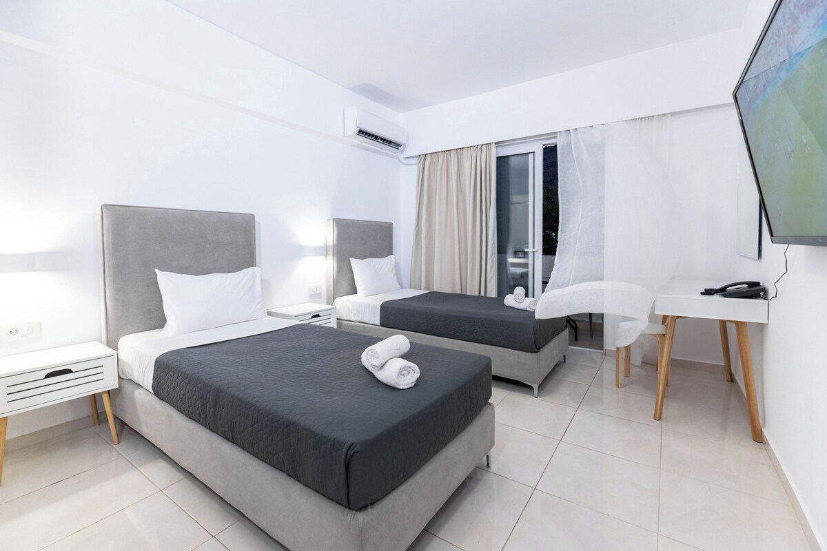 A hotel room with two beds, a desk, a chair, a TV, and an air conditioning unit.