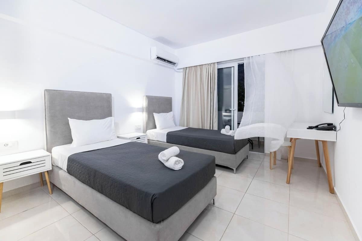 A hotel room with two beds, a desk, a chair, a TV, and an air conditioning unit.