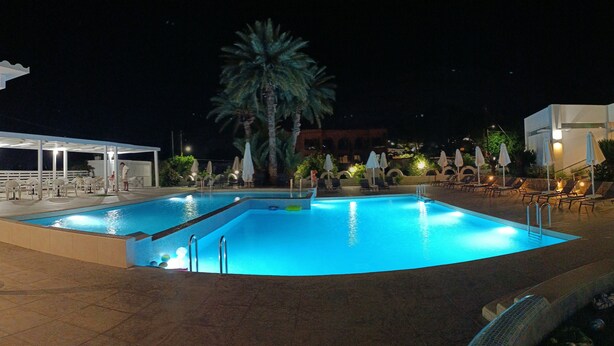 A nighttime swimming pool with lounge chairs and umbrellas, surrounded by palm trees and a building in the background. - Pefkos Breeze Family Resort & Spa (Rhodes)