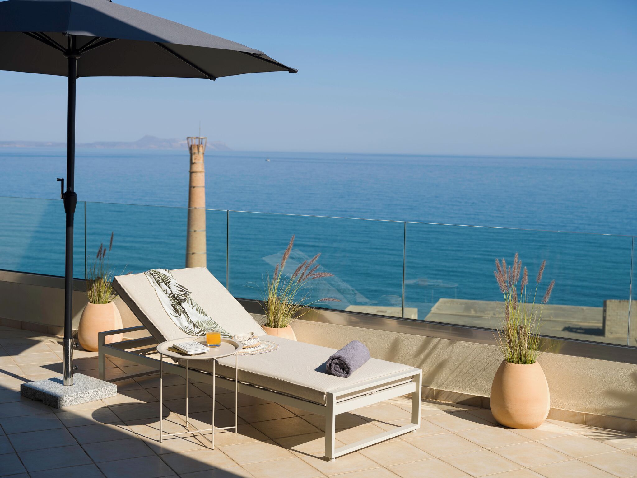 A sun lounger with a view of the sea, a potted plant, and a table with a book and a glass of orange juice.
