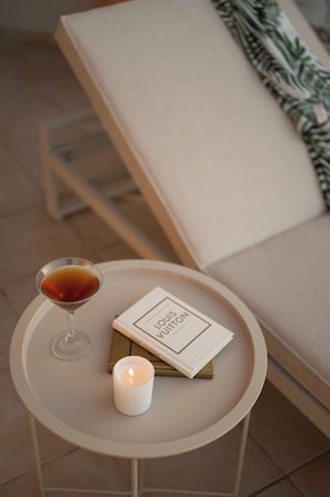 Ein runder, weißer Tisch mit einem Martini-Glas, einer Kerze und einem Buch mit dem Titel 'LOUIS VUITTON' vor einer beigen Couch im Hintergrund.