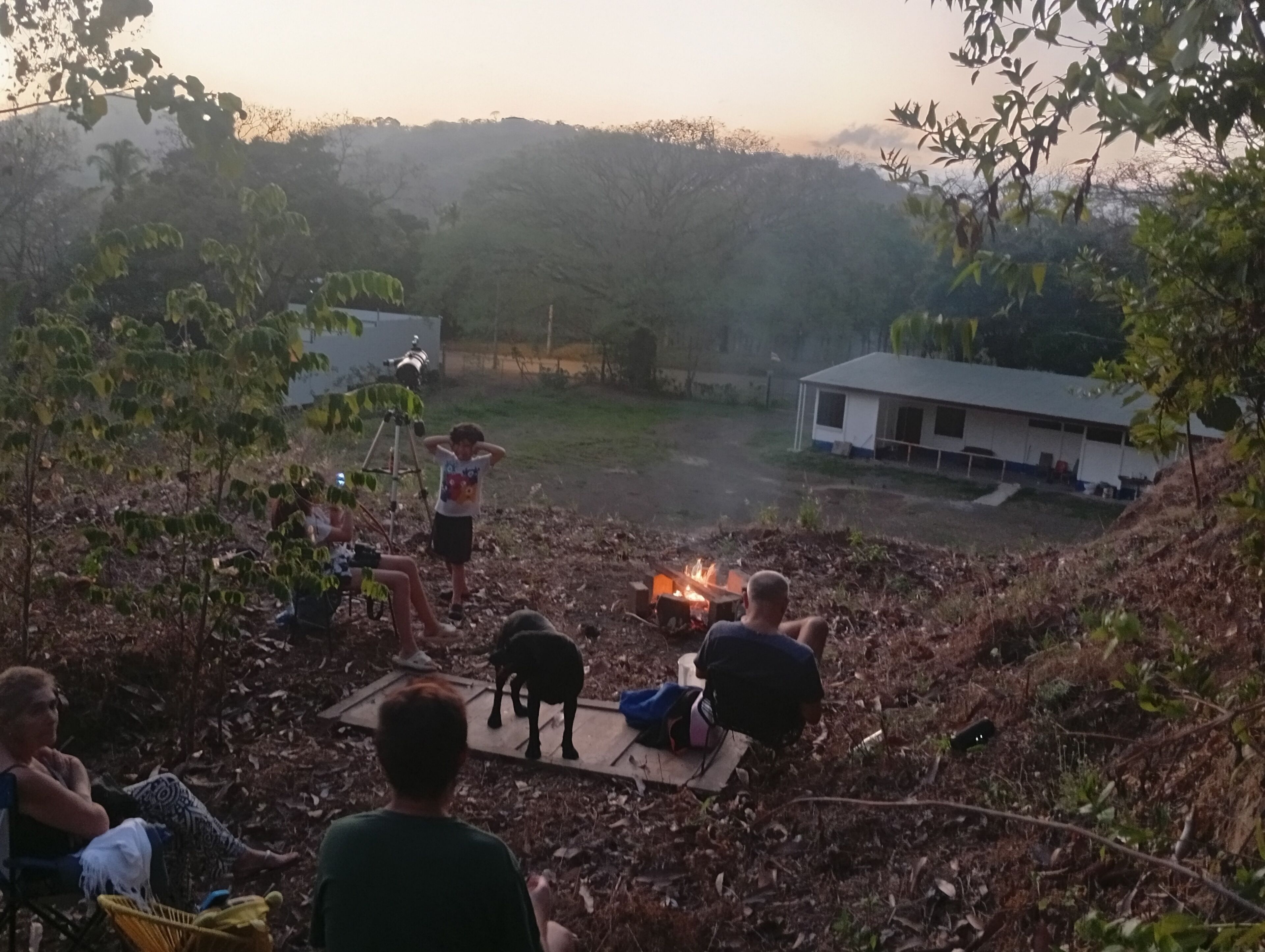 Een groep mensen zit rond een kampvuur in een natuurlijke omgeving, met bomen en een gebouw op de achtergrond.
