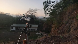 Un telescopio sobre un trípode con una lata de bebida cerca, un edificio al fondo y un fuego en primer plano.